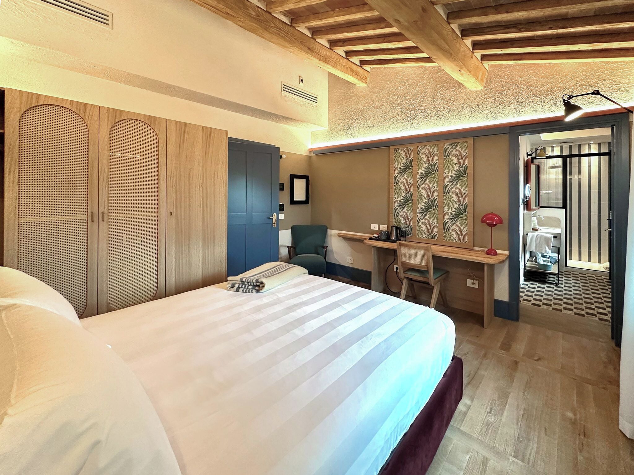 A hotel room with a large bed, a desk, a chair, a wardrobe, and a bathroom visible through an open door.