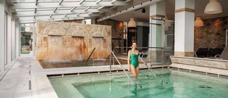 A woman in a green swimsuit standing in a pool with a stone wall and a glass ceiling.