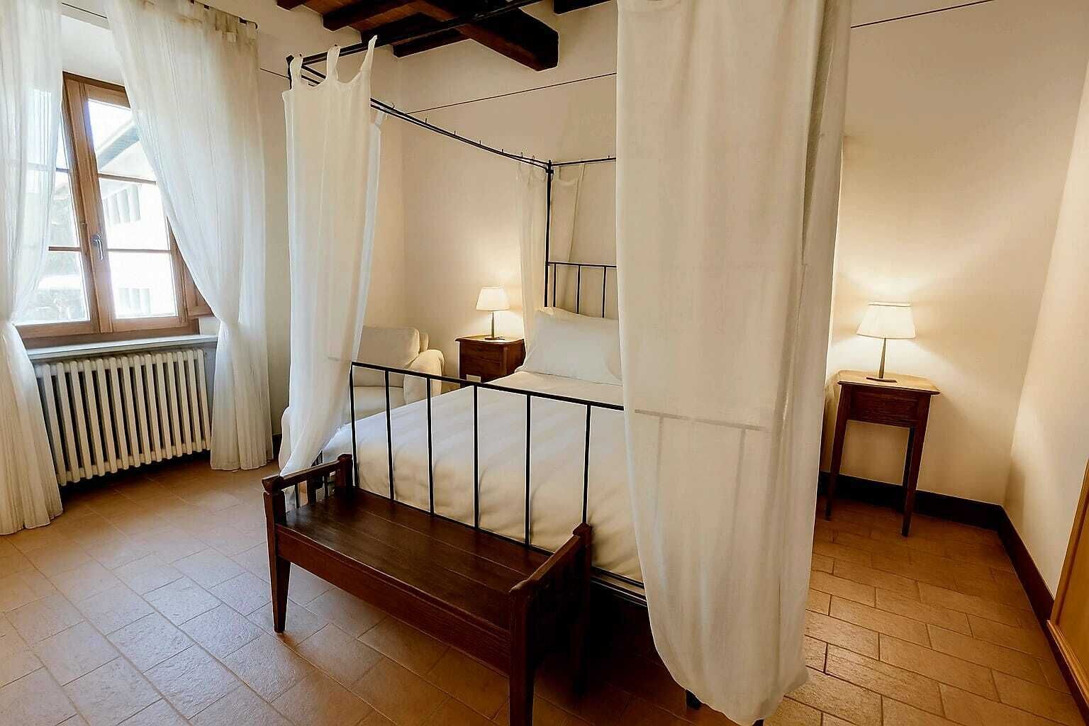 A bedroom with a four-poster bed, a wooden bench, two bedside tables with lamps, a radiator, and a window with curtains.