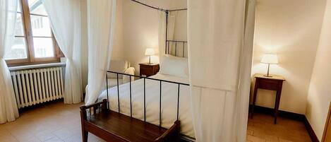 A bedroom with a four-poster bed, a wooden bench, two bedside tables with lamps, a radiator, and a window with curtains.