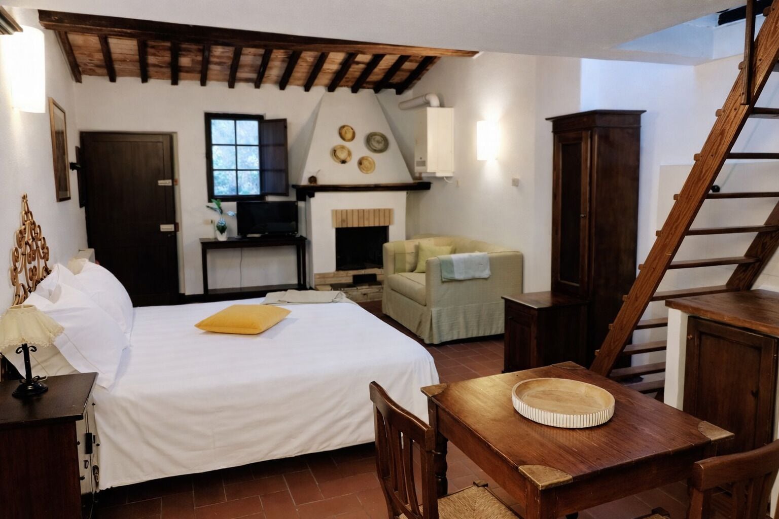 A bedroom with a bed, a wooden table, a chair, a sofa, a fireplace, a television, and a staircase.