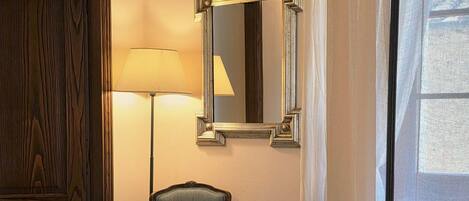 A room with a wooden door, a chair, a lamp, a mirror, and a window with sheer curtains.
