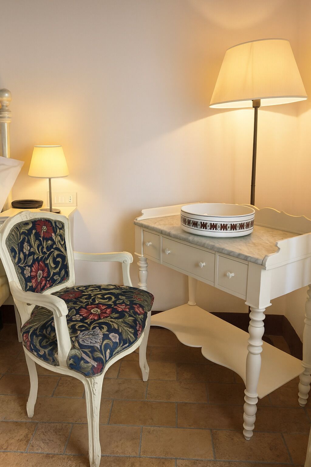 Een kamer met een wit bureau, een stoel met bloemenmotief bekleding, een lamp en een schaal op het bureau.