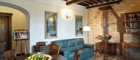 A cozy living room with a blue leather sofa, wooden coffee table, and a dining table with a basket of apples. There is a bookshelf filled with books and a lamp on a side table.