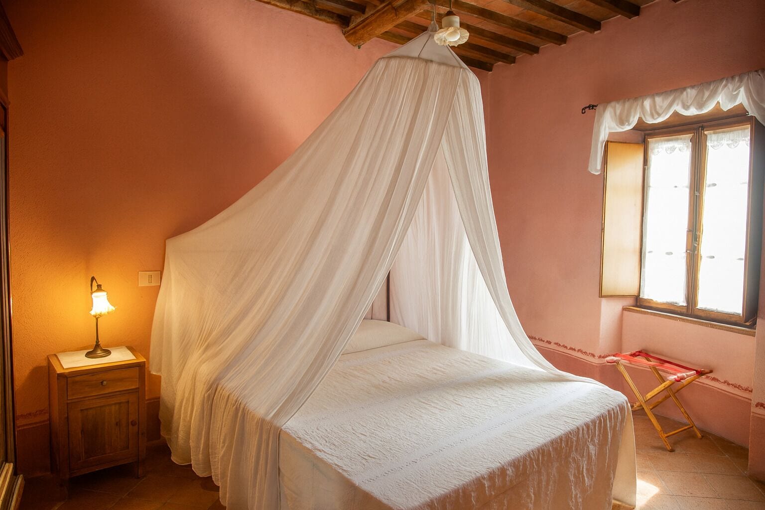 A bedroom with a canopy bed, a bedside lamp, a window with curtains, and a small table with a chair.