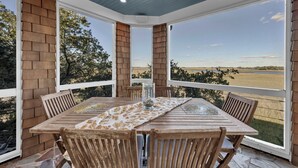A covered patio with a wooden table and chairs, large windows, and a view of a field. - Beauitful marsh front, six bedroom home with infinity pool with hot tub. (Bald Head Island)