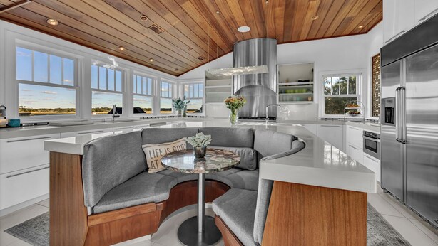 A modern kitchen with a curved island, built-in seating, and large windows offering a view of the landscape. - Beauitful marsh front, six bedroom home with infinity pool with hot tub. (Bald Head Island)