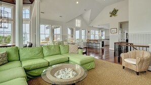 A spacious living room with a green sectional sofa, a round coffee table, and a wicker chair. - Five bedroom home on Bald Head Island's East Beach, make your next island getawa (Bald Head Island)