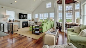 A spacious living room with a fireplace, green sofas, a coffee table, and a large window overlooking a scenic view. - Five bedroom home on Bald Head Island's East Beach, make your next island getawa (Bald Head Island)