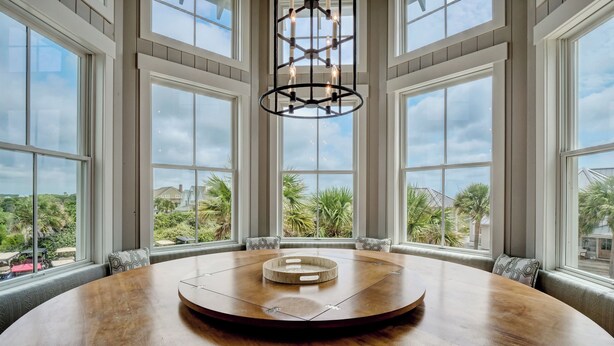 A round wooden table with a central wooden piece, surrounded by chairs with patterned cushions, in a room with large windows and a hanging light fixture. - Five bedroom home on Bald Head Island's East Beach, make your next island getawa (Bald Head Island)