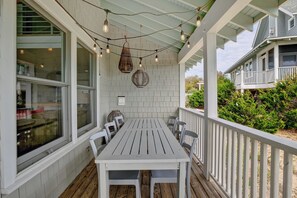 A covered outdoor dining area with a white table and chairs, string lights, and a view of the surrounding greenery. - Renovated Ocean View Home, 3 Bedrooms Bald Head Island, NC (Bald Head Island)