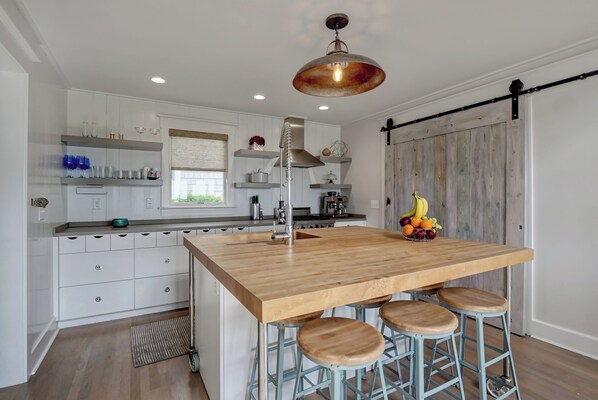 Une cuisine moderne avec des meubles blancs, un îlot central en bois doté de tabourets, un luminaire suspendu et un panier de fruits sur l’îlot.