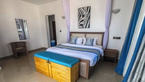 A bedroom with a bed, a wooden chest, a nightstand, a mirror, and a painting on the wall. - PV 102 (Kibaoni)