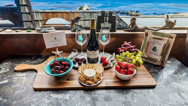 A table with a bottle of wine, glasses, a starfish decoration, and various fruits and cheeses. - 3965 Marine Gardens - Oceanfront with Bay Views (Pacific Grove)
