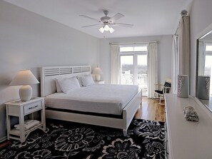A bedroom with a bed, nightstands, a ceiling fan, a window with curtains, and a view of a balcony. - Oceanfront Beach Cottage with Direct Beach Access (Bald Head Island)