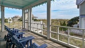 A covered outdoor area with wooden benches and large windows overlooking a beach and houses. - Oceanfront, 4 Bedrooms, BHI (Bald Head Island)