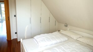 A bedroom with a sloped ceiling, white bedding, and a wardrobe with silver handles. - Ferienhaus Sturmfrei auf Fehmarn (Fehmarn)