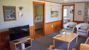 Una camera d'albergo con televisione, un tavolo in legno, sedie in vimini, un letto e un quadro raffigurante una spiaggia.