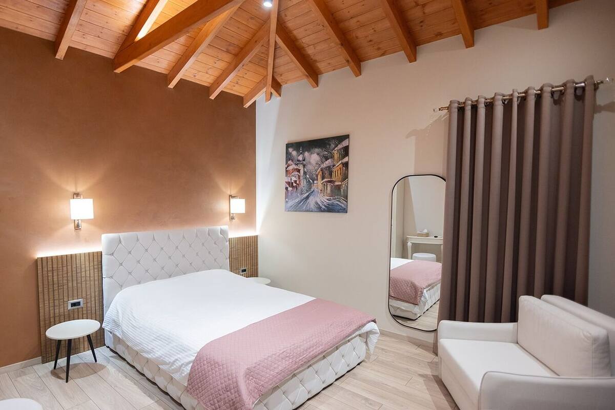 Un dormitorio con cama, mesitas de noche, una silla, un espejo y un cuadro en la pared.