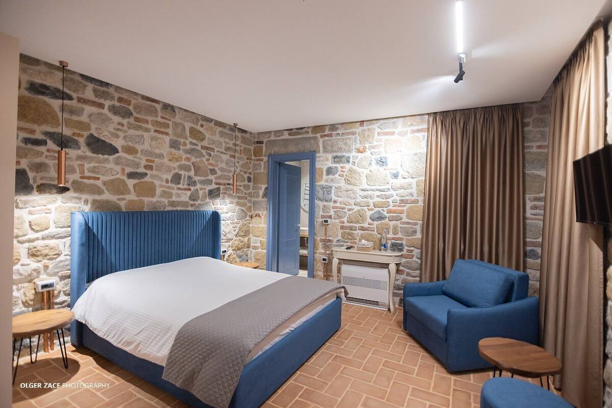 Un dormitorio con pared de piedra, cama con cabecera azul, sillón azul, una mesita pequeña y un televisor montado en la pared.