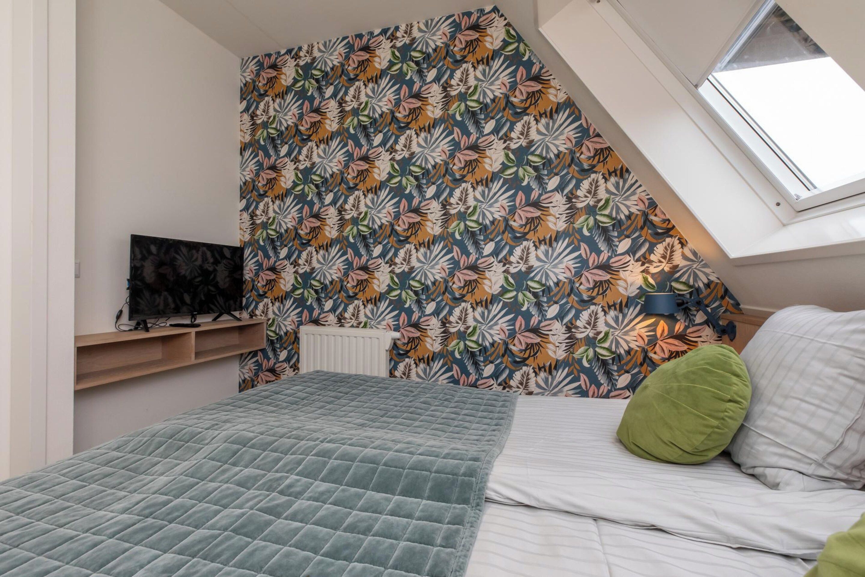 Une chambre avec un plafond en pente, un lit recouvert d’une housse matelassée, un oreiller vert, un téléviseur fixé au mur et un radiateur.