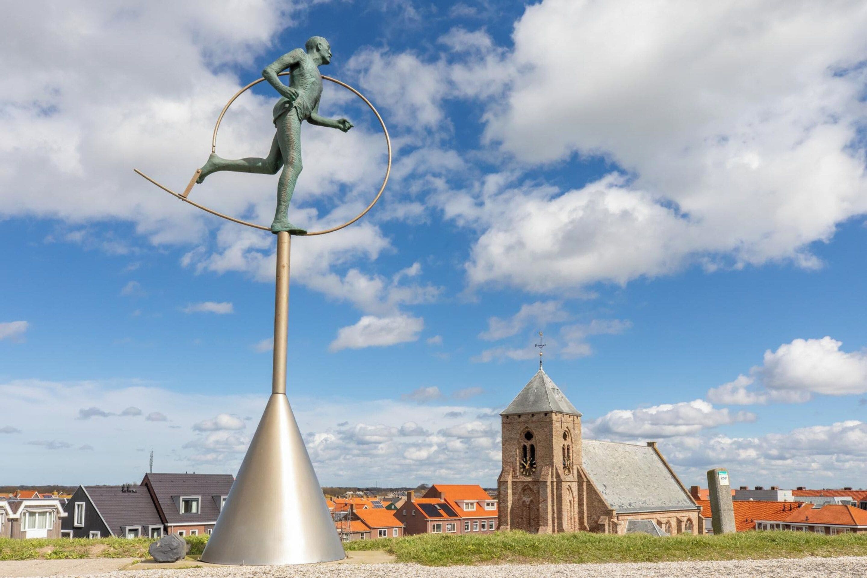 Una estatua de una figura equilibrándose sobre un poste con un aro circular, con un fondo que muestra una iglesia y edificios residenciales.