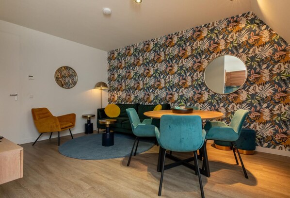 A modern living room with a round wooden table, teal chairs, a green sofa, and a floral-patterned wallpaper. - Apartment in Zoutelande Near the Beach (Zoutelande)