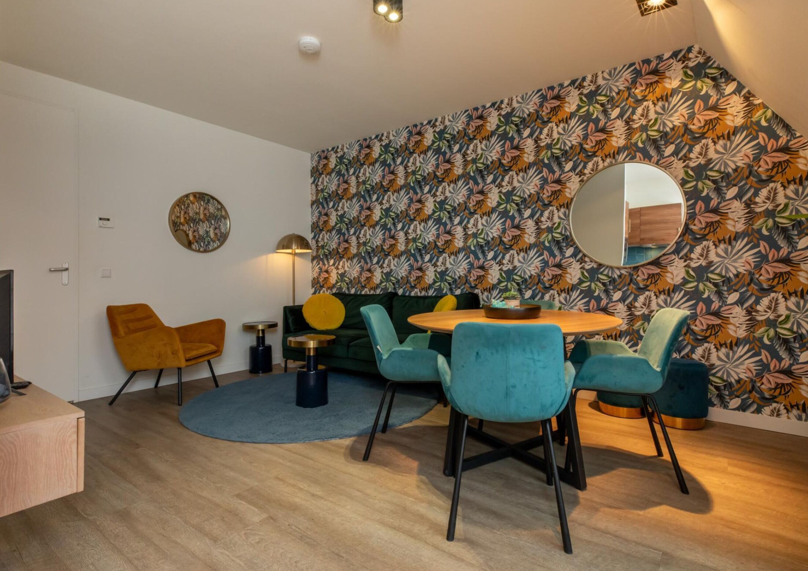 A modern living room with a round wooden table, teal chairs, a green sofa, and a floral-patterned wall.