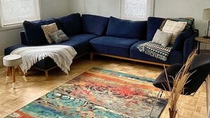 A corner living room with a dark blue sectional sofa, a patterned rug, and two windows with blinds.