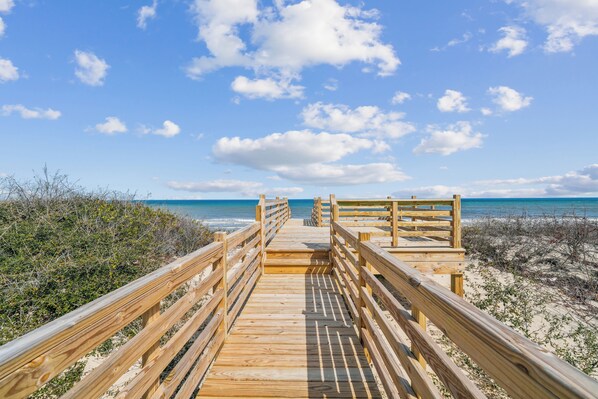 A wooden boardwalk leading to a beach with a clear sky and ocean in the background.