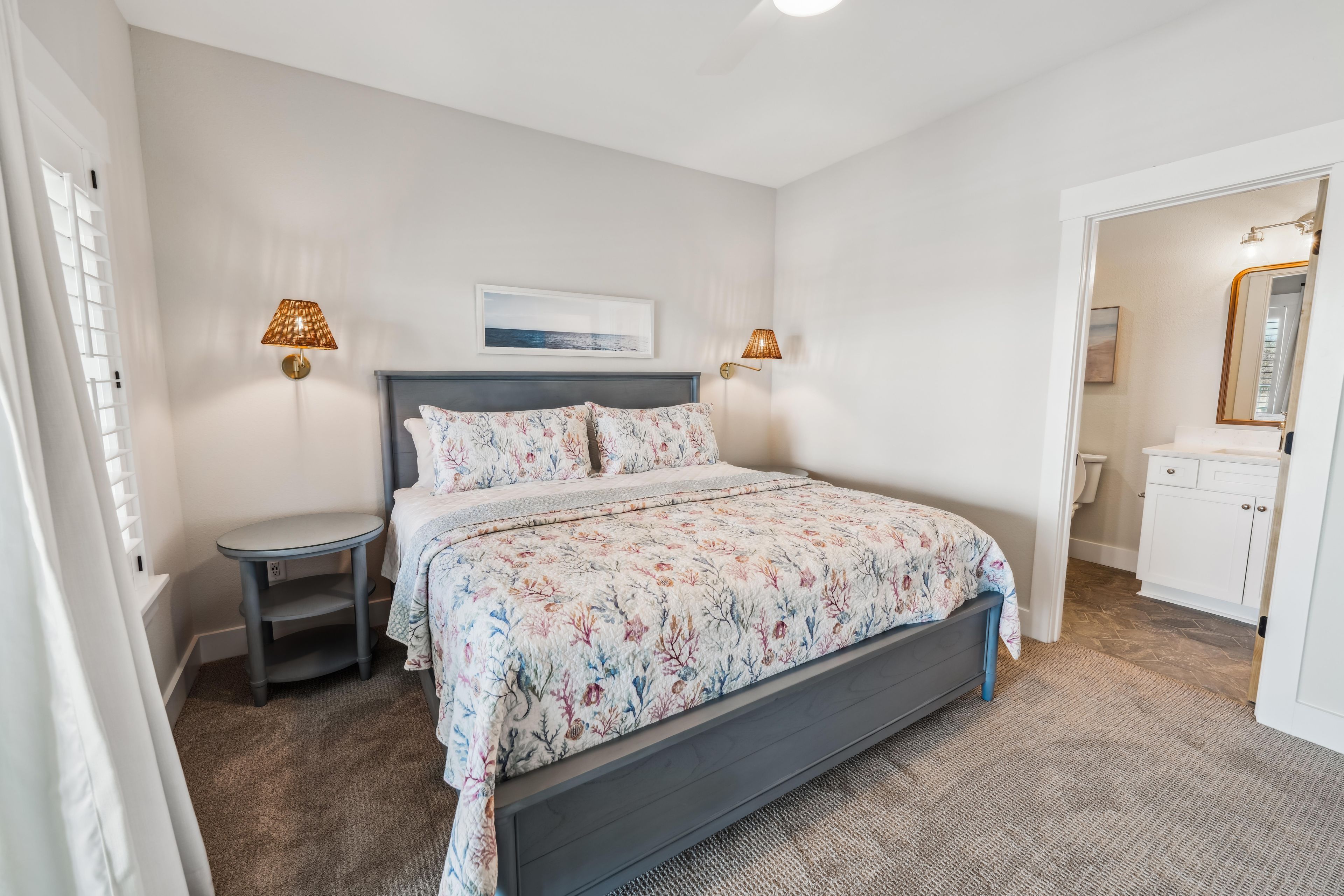A bedroom with a bed, bedside tables, a nightstand, a wall-mounted lamp, a framed picture, and a bathroom visible through an open door.