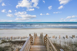 Un paseo marítimo de madera que conduce a una playa arenosa, con un cielo azul despejado y el océano al fondo.