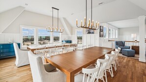 A spacious dining area with a long wooden table and white chairs, large windows with a view of the landscape, and a modern chandelier. - Daniels -- Brand New Luxury Oceanfront Home (Corolla)