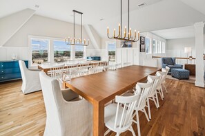 A spacious dining area with a long wooden table and white chairs, large windows with a view of the landscape, and a modern chandelier.