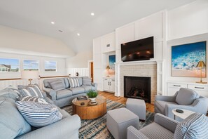 Un salón espacioso con chimenea, un televisor de pantalla plana grande y vistas al exterior a través de las ventanas.