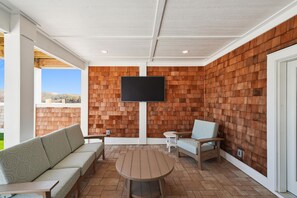 Een overdekt terras met een bakstenen muur, een flatscreen televisie boven een ronde tafel, en zitplaatsen, waaronder een bank en twee stoelen.