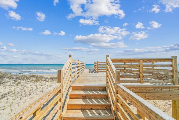 A wooden boardwalk leading to the beach with a clear sky and ocean in the background.