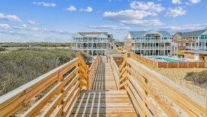 A wooden boardwalk leading to a beachfront property with multiple buildings, a pool, and a clear sky. - Owens -- Brand New 12 BR Luxury Oceanfront (Corolla)