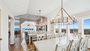 A spacious dining area with a large white table and chairs, a modern chandelier, and a view of the ocean through large windows. - Owens -- Brand New 12 BR Luxury Oceanfront (Corolla)