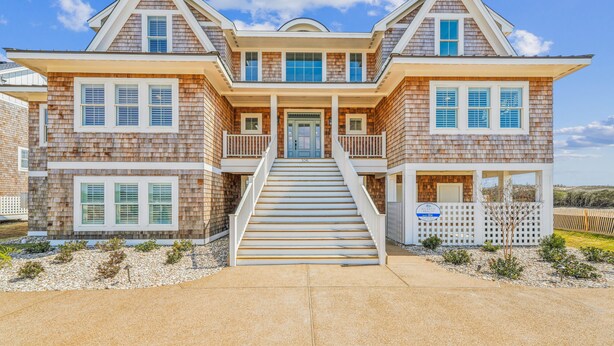 A two-story house with a front porch, a staircase leading to the entrance, and a small garden area with gravel and plants. - Owens -- Brand New 12 BR Luxury Oceanfront (Corolla)