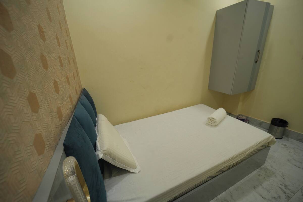 A single bed with a white mattress, a rolled-up white towel, and a blue pillow. A grey cabinet is mounted on the wall to the right of the bed.