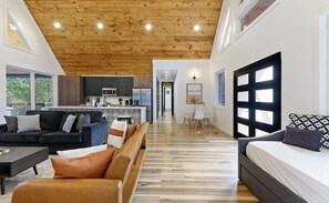 Un intérieur moderne avec un plafond en bois, une cuisine dotée de meubles sombres, un coin repas avec une table et des chaises blanches, et un grand lit agrémenté d’oreillers à motifs.