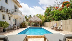 Piscina com espreguiçadeiras, um gazebo de palha e um prédio branco com aparelhos de ar condicionado.