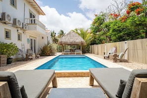 A swimming pool with lounge chairs, a thatched gazebo, and a white building with air conditioning units.