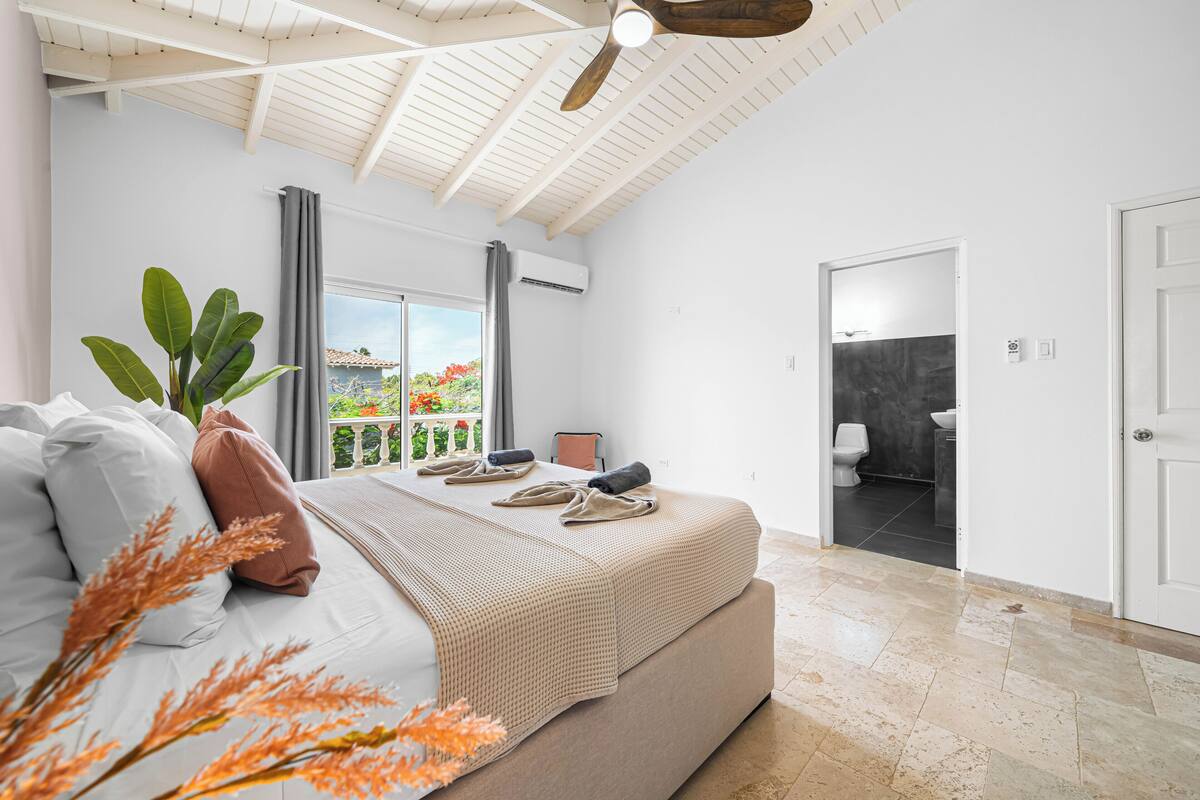 A bedroom with a bed, a ceiling fan, a window with curtains, and a view of the outdoors.