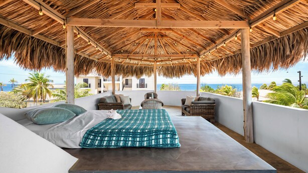 Un dormitorio con techo de paja, una cama, almohadas y vista al océano.