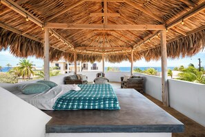 Un dormitorio con techo de paja, una cama, almohadas y vista al océano.