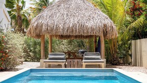 Una piscina con estructura de techo de paja, dos sillas de descanso y una mesa de madera.