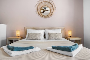 Uma cama bem arrumada com colcha estampada, duas mesas de cabeceira com luminárias e um relógio de parede.