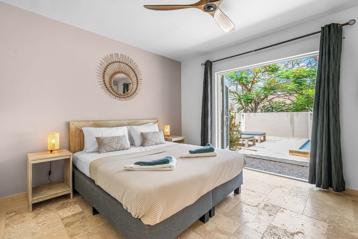 A bedroom with a bed, bedside tables, a ceiling fan, a window with curtains, and a view of the outside.